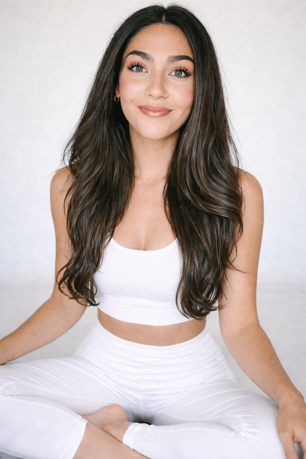 Smiling woman with long dark hair in a white sports bra and leggings, sitting cross-legged against a light background.