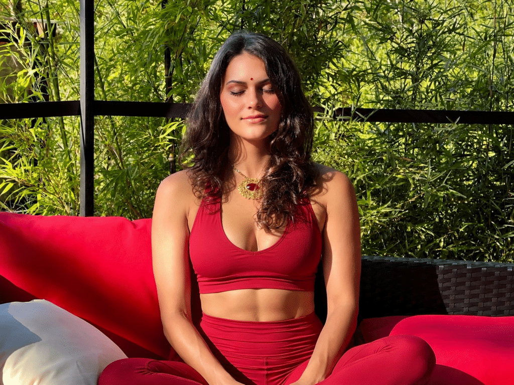 Woman in red athletic wear sits cross-legged on a red outdoor couch, eyes closed in meditation with green plants in the background.