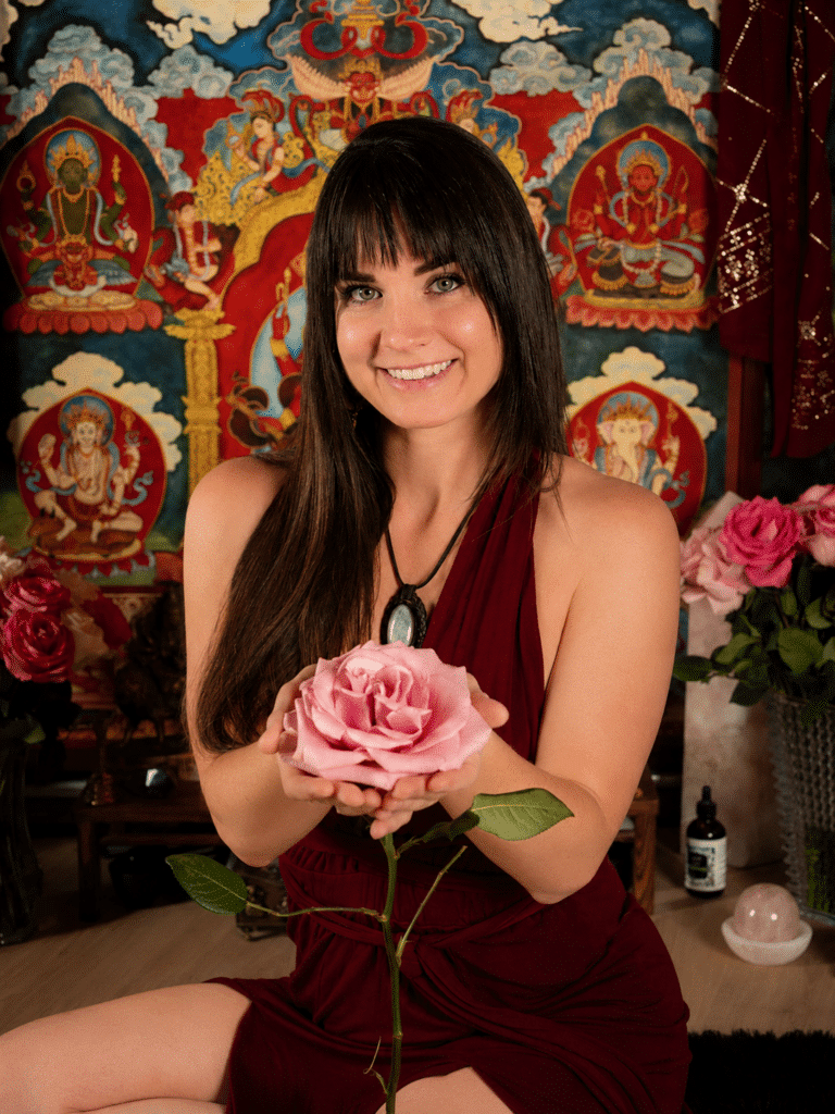 Tantra for Coupkes - Temple of Bliss Smiling woman in a burgundy dress sits and offers a pink rose toward the viewer, with a colorful tapestry backdrop.