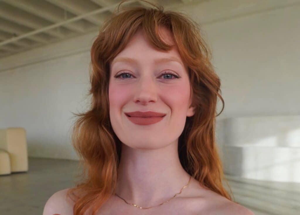 Los Angeles Tantra Massage - Temple of Bliss Close-up of a smiling red-haired woman wearing a delicate gold necklace in a softly lit indoor setting.