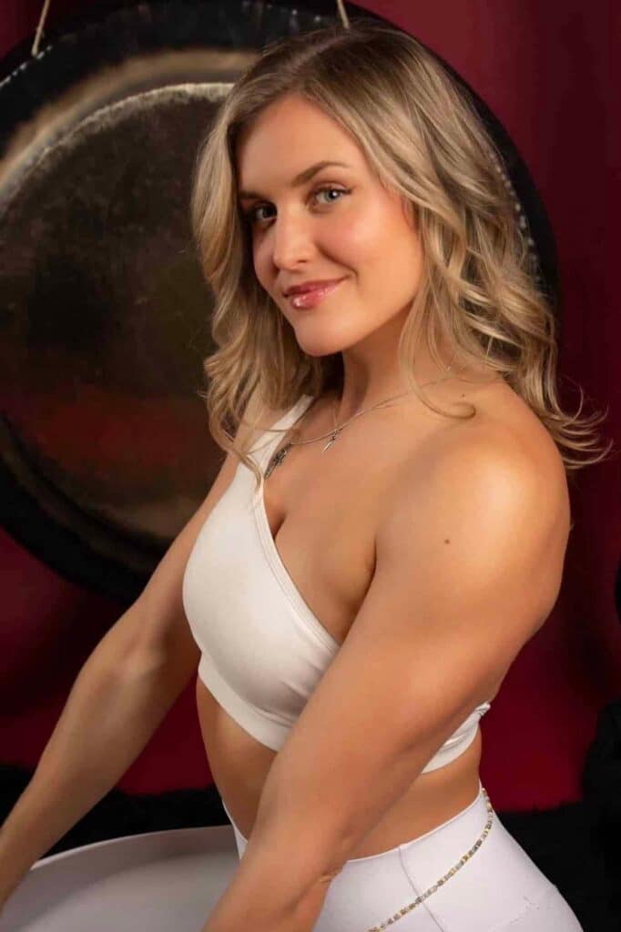 San Francisco Tantra Massage - Temple of Bliss Portrait of a smiling woman with blonde hair wearing a white sports bra, posing beside a large gong against a dark red backdrop.